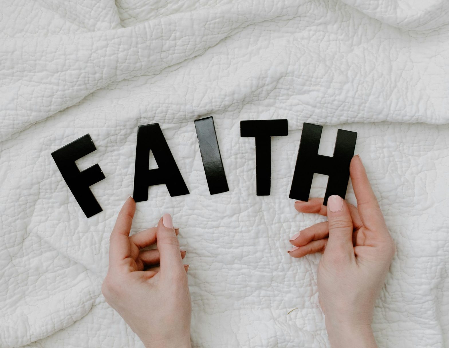 Two hands against a white terry cloth background. Hands pointing toward black vinyl letters spelling the word FAITH in all capital letters.